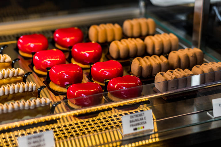 Sobremesas dispostas em vitrine fria, algumas parecendo bombas de chocolate, outras corações vermelhos brilhantes e outras uma espécie de ziguezague de chocolate