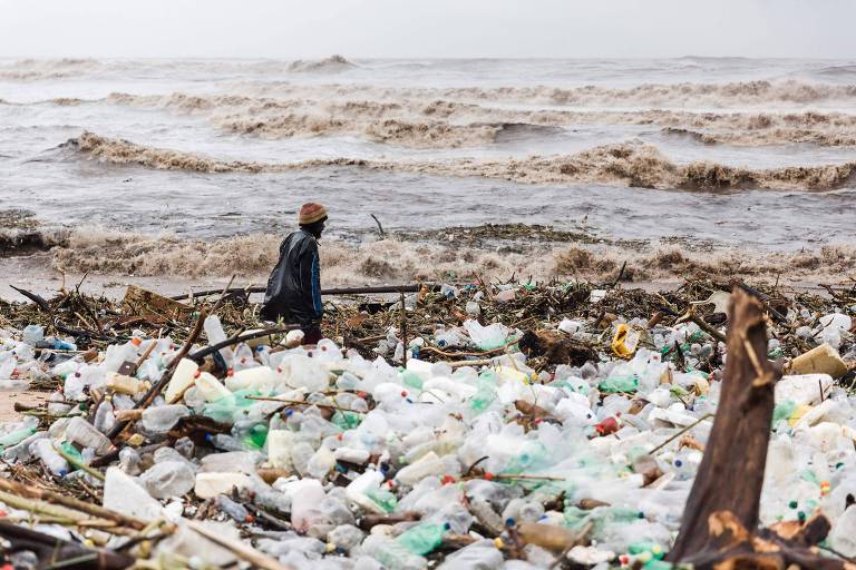 Homem está ao lado de muitos pedaços de plástico no que  parece ser uma praia ou o leito de um rio. Ao fundo, a água está revolta, formando ondas de cor marrom