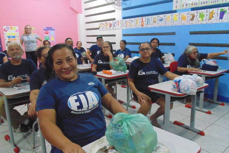 Sala de aula do EJA (Ensino de Jovens e Adultos) com ao menos 14 alunos sentados, nas carteiras, vestindo camisa azul com a inscrição EJA. Ao fundo, no alto da parede, folhas de sulfites com letras inscritas usadas para ensinar o alfabeto