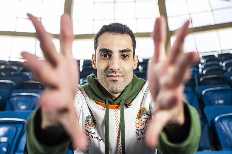 retrato de Douglas Souza, atleta de vôlei, sentado em arquibancada com mãos abertas em primeiro plano