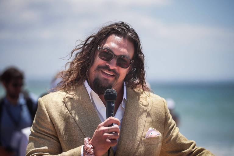 Jason Momoa usa casaco bege, camisa branca e óculos escuros e segura microfone