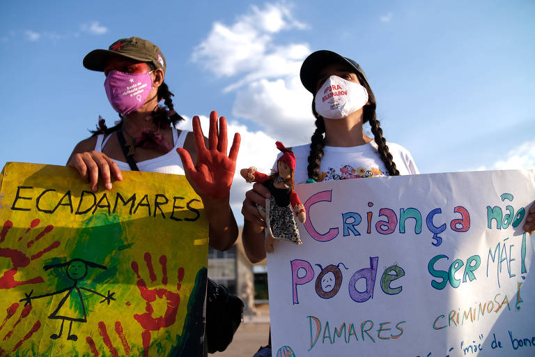 Duas ativistas engajadas em uma manifestação. Uma das faixas diz que criança não pode ser mãe. Elas estão ao ar livre, com expressões sérias e determinadas, usando máscaras de proteção