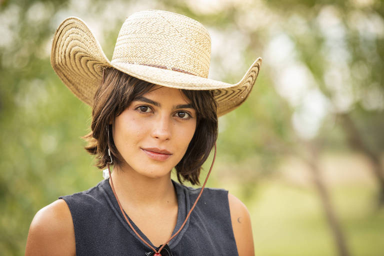 Em foto colorida, mulher de chapéu olha para câmera e sorri levamente