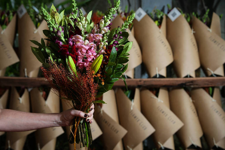 Uma mão segura um buquê de flores. Ao fundo, há diversos outros buquês.