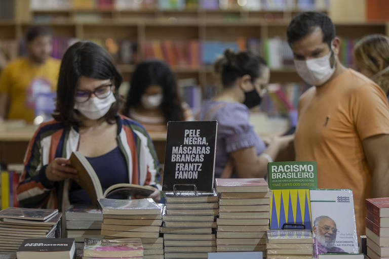 Imagem mostra menina manuseando um livro que está sobre uma pilha de outros livros. Outras pessoas estão atrás dela, num estande.