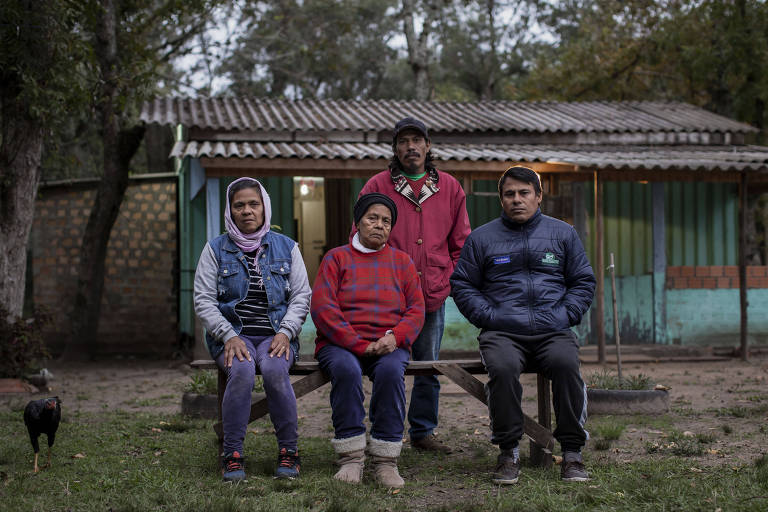 Dois homens e duas mulheres com traços indígenas olham para a câmera sentados ou em pé em frente a uma casa. Eles vestem casacos de frio