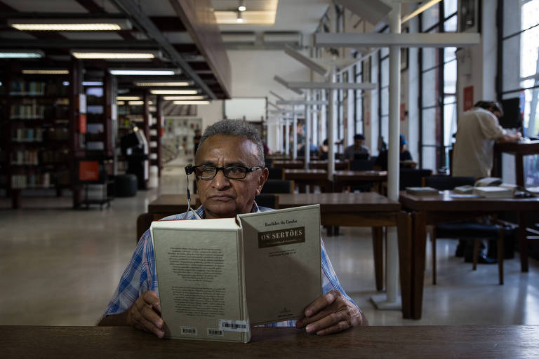 Homem usa óculos pretos com um aparelho acoplado do lado direito, com o qual ele mira para o livro "Os Sertões", que ele mantém aberto em mesa de biblioteca