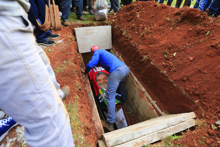 Imagem mostra homem colocando toalha vermelha com rosto de Lula em cima do caixão de Marcelo de Arruda, dentro de uma cova. O buraco está cercado por terra, e é possível ver as pernas de amigos e familiares que compareceram ao sepultamento.