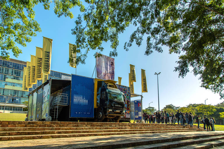 Foto mostra a estrutura do caminhão-museu. Ao lado dele, estão painéis com pinturas. Acima, algumas faixas amarelas trazem palavras relacionadas. Pessoas acessam a lateral do veículo para visitar a exposição