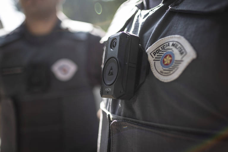foto mostra câmera corporal acoplada a farda de policial. é possível ver, o símbolo da polícia militar sobre a farda cinza
