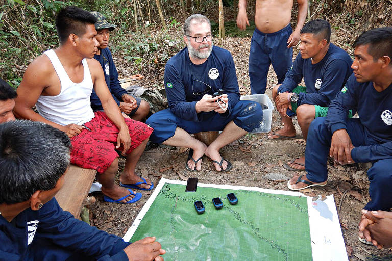 Em uma roda de pessoas em um local ao ar livre, Bruno Pereira segura um aparelho que parece ser um rádio, com uma pequena antena, enquanto os demais escutam; ao centro, sob um mapa impresso disposto no chão, há alguns aparelhos de celular