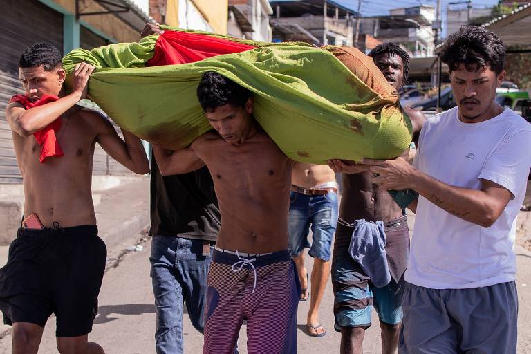 Três homens, dois pardos e um branco, carregam um corpo enrolado em um lençol verde em uma via do Complexo do Alemão, no Rio de Janeiro 