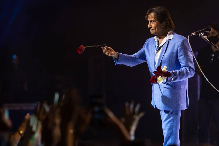 De cima de um palco, homem de terno azul segura rosa vermelha em direção ao público
