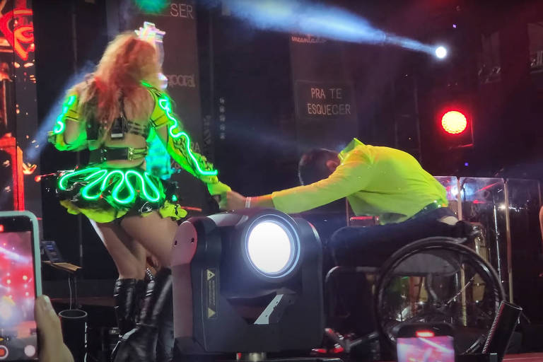 A cantora Joelma, usando roupas verde neon (saia e maiô), dá a mão, em um passo de dança, para um bailarino que está em uma cadeira de rodas. Ele usa camisa amarelo neon e calça preta e está com o corpo inclinado para frente
