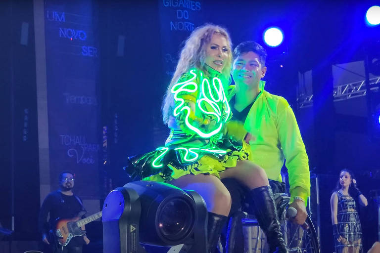 A cantora Joelma, com roupas fosforescente, senta-se no colo de um bailarino cadeirante, com camisa amarelo neon.