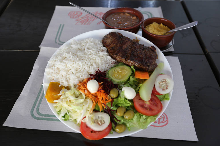 Imagem mostra um prato feito, acompanhado de duas tijelas com caldo de feijão e farofa. No prato, há arroz branco, salada com muitas verduras e legumes, como alface, cenoura, pepino, repolho, tomate, chuchu e azeitonas, além de duas proteínas, carne bovina e ovo de codorna.