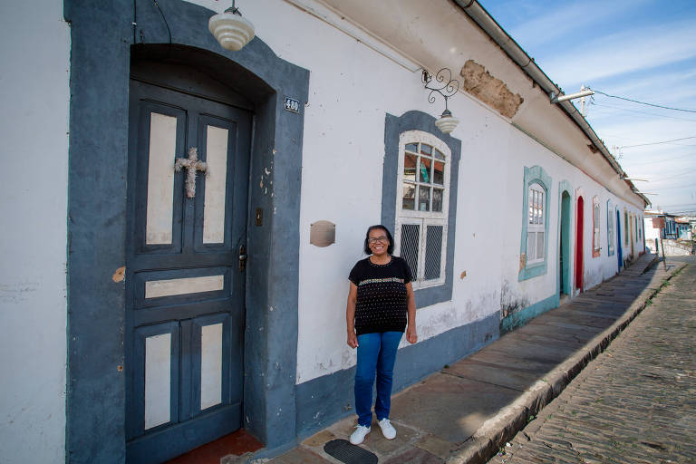 Mulher de calça jeans e blusa preta posa em frente à casa histórica de porta cinza; ao fundo, há casas com portas coloridas em azul e vermelho