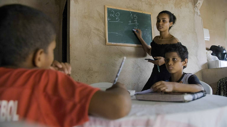 Duas crianças estão sentadas, estudante, e uma moça de pé escreve numa pequena lousa.