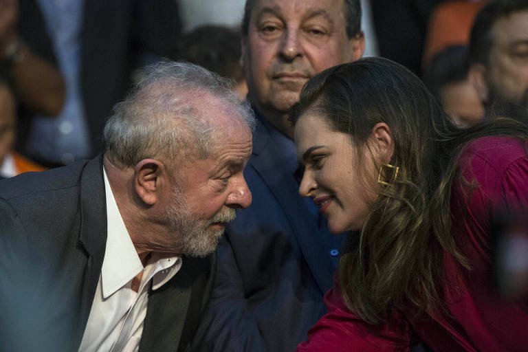 Na foto, Lula está à esquerda, de terno preto e camisa branca, e Marília Arraes está de vermelho, à direita. Lula fala e Arraes ouve, os dois inclinando-se para ficar mais próximos. Atrás, participantes da convenção, um deles, ao centro da foto, observando a conversa entre os dois