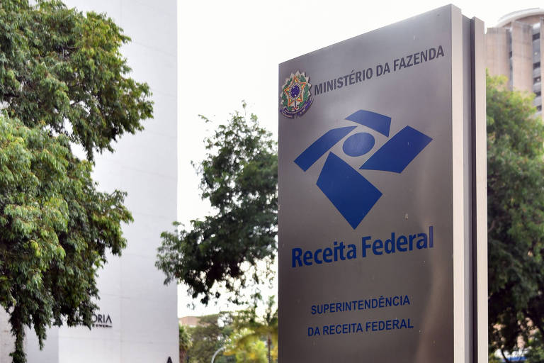 A imagem mostra um totem de sinalização da Receita Federal do Brasil, com o logotipo e nome da instituição em destaque. O totem está localizado ao lado de uma calçada, com árvores ao fundo.
