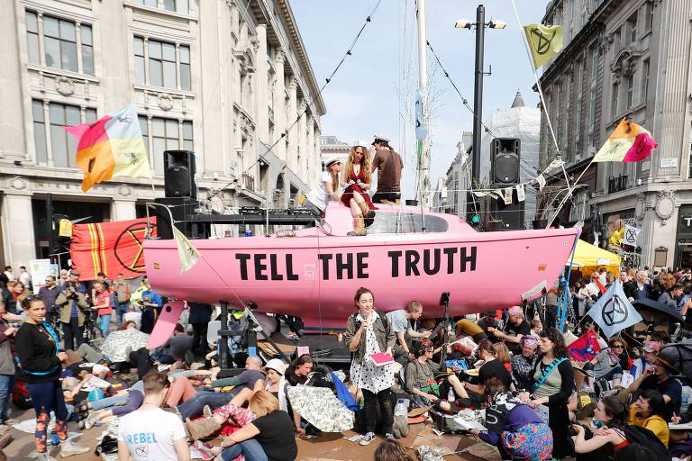 Pessoas em volta de um barco cor de rosa colocado em uma rua; nele está escrito "tell the truth" (conte a verdade)