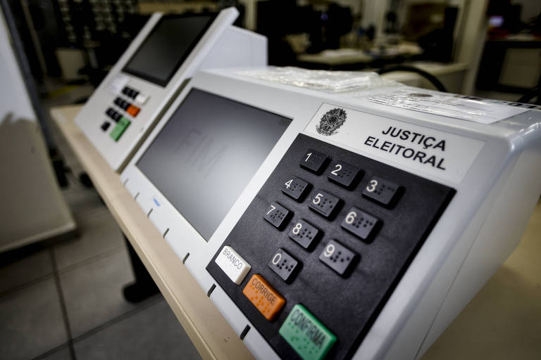 Urna eletrônica é branca com teclado preto e três teclas coloridas, verde, vermelha e branca. Elas estão sobre mesa