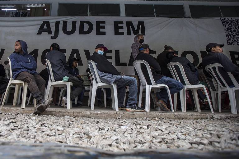 A imagem mostra um grupo de pessoas sentadas em cadeiras plásticas brancas, aguardando em um local que parece ser um centro de ajuda. Ao fundo, há uma grande faixa com a inscrição 'AJUDE EM FAVELAS'. O ambiente é simples, com piso de pedras soltas e uma parede ao fundo. Algumas pessoas estão usando máscaras, e a maioria está vestida com roupas casuais e agasalhos.