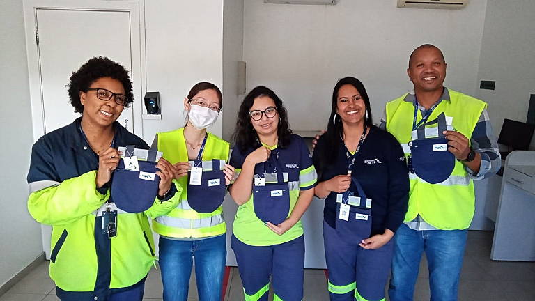 Imagem colorida mostra quatro mulheres, da esquerda pra a direita, e um homem; eles seguram sacos de lixo de pano para automóveis na cor azul.