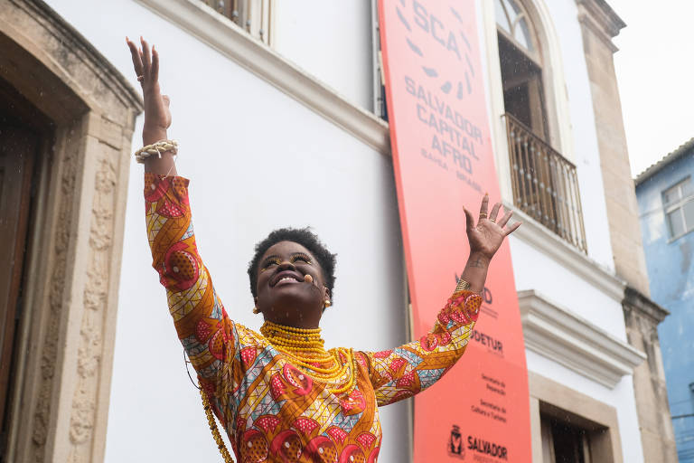 Imagem colorida mostra uma mulher negra, do tórax para cima, com os dois braços levantados; ela tem cabelo curto e veste uma blusa de manga comprida em tons de amarelo e laranja; ao fundo tem uma parede branca, de um imóvel antigo.