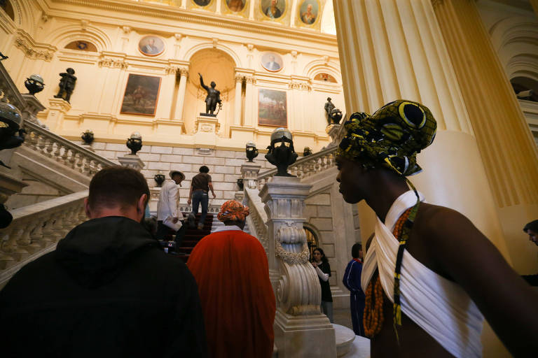 garota negra está em primeiro plano, de costas, seguindo um grupo que sobe as escadas do saguão de entrada do museu