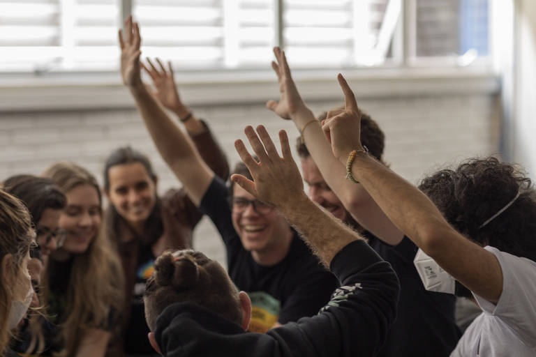 A imagem mostra um grupo de pessoas em um ambiente interno, aparentemente celebrando ou participando de uma atividade em grupo. Todos estão sorrindo e levantando as mãos, demonstrando entusiasmo e alegria. O fundo é desfocado, mas parece ser uma sala com janelas e paredes de tijolos.