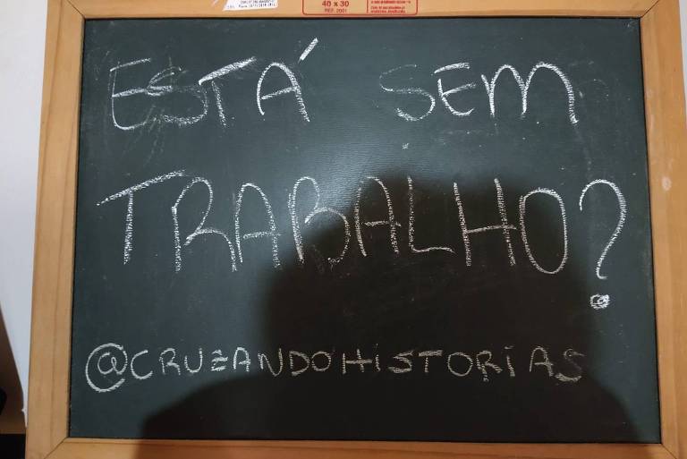 Retrato de lousa com a inscrição, em giz branco sobre o quadro verde, que pergunta: "Está Sem Trabalho?". A lousa ainda traz o endereço eletrônico da ONG Cruzando Histórias, liderada por Bia Diniz, o @cruzandohistórias