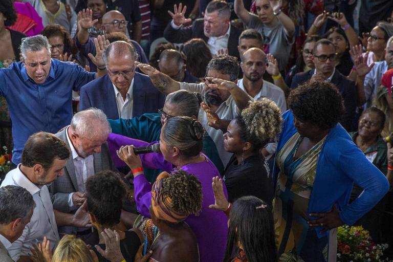 Um aglomerado de pessoas reunidas em volta de Luiz Inácio Lula da Silva e Geraldo Alckmin estão com as mãos estendidas orando por ele.