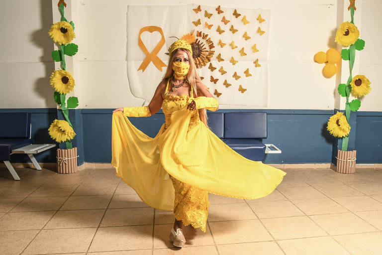 Médica colombiana usa vestido com uma saia como véu (que ela segura nas mãos), máscara e uma coroa, tudo amarelo; o fundo tem três girassóis falsos à esquerda e outros três à direita, colados na parede