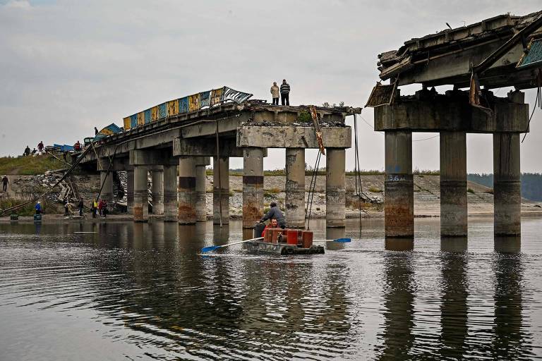 Moradores de Stari Saltiv, em Kharkiv, atravessam de barco o rio Donets, sob ponte destruída na guerra