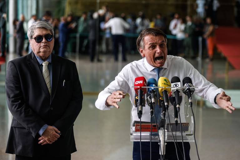 Presidente Jair Bolsonaro durante entrevista no Palácio do Alvorada. Ele é um homem branco, que veste uma camisa branca. Ele está visivelmente irritado, gesticulando bastante. Ao seu lado, está o apresentador José Luiz Datena, que veste um terno preto e usa óculos escuros.