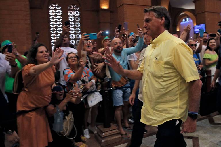 Fotografia colorida mostra o presidente do Brasil, Jair Bolsonaro, em primeiro plano. Ele veste uma camiseta social na cor amarela. Está com o braço direito levantado cumprimentando apoiadores que acenam para ele. 