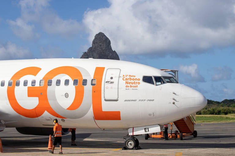 Fotografia colorida da parte da frente de um avião Gol, visto de perfil virado para a direita e que informa com um adesivo posicionado logo antes da janela dos pilotos A 1° rota carbono neutro do Brasil: Recife a Noronha. O avião é da cor branca e tem o logo da Gol estampado na lateral, na cor laranja. Um funcionário do aeroporto é visto à distância no canto inferior esquerdo, usando um colete de segurança e protetor de ouvido da cor laranja