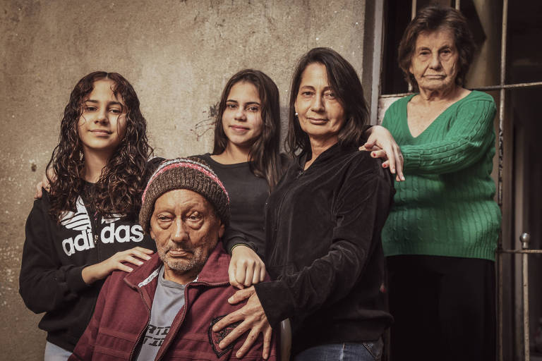 Imagem colorida mostra, da cintura para cima, duas jovens à esquerda, outras duas mulheres à direita, e um homem idoso, sentado à frente; As jovens e uma das mulheres vestem blusas de moletom na cor preta; a mulher à direita veste blusa verde. O homem está com um gorro marrom e uma camisa vermelha.