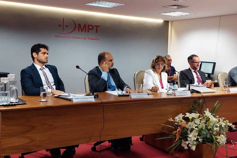 Na mesa do evento os participantes são três homens e uma mulher, eles estão sentados, no fundo vemos a logomarca do Ministério Público do Trabalho