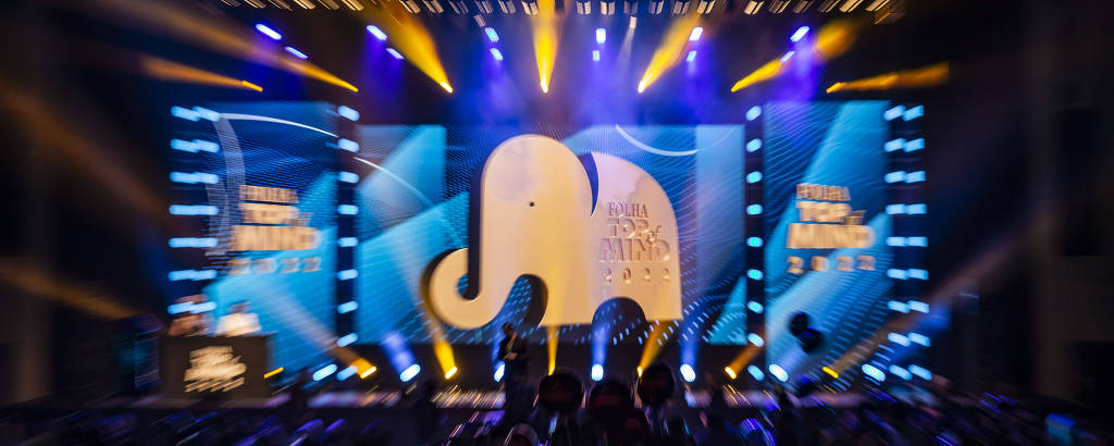 Fotografia colorida do palco da cerimônia da Folha Top of Mind no Tokio Marine Hall; há um telão com projeção de uma ilustração de um elefante branco, símbolo da premiação, e se vê escrito também Folha Top of Mind 2022; em um fundo azul; na parte inferior da imagem, bem escura, há a plateia