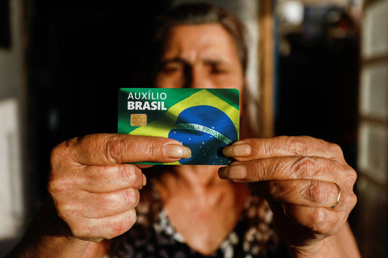 Mulher mais velha segura, em frente ao rosto, cartão do Auxílio Brasil