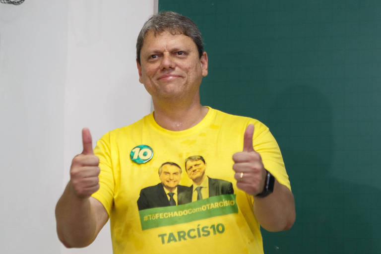 Homem de cabelo curto e camiseta amarelo ouro com foto dele e de bolsonaro faz sinal de positivo com os dois polegares em frente a lousa verde e parede branca