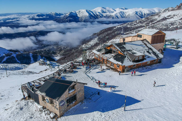 Vista aérea de um resort de esqui com edifícios robustos aninhados na neve branca, cercados por pistas de esqui com visitantes esquiando em um dia ensolarado. Ao fundo, montanhas cobertas de neve se erguem acima das nuvens