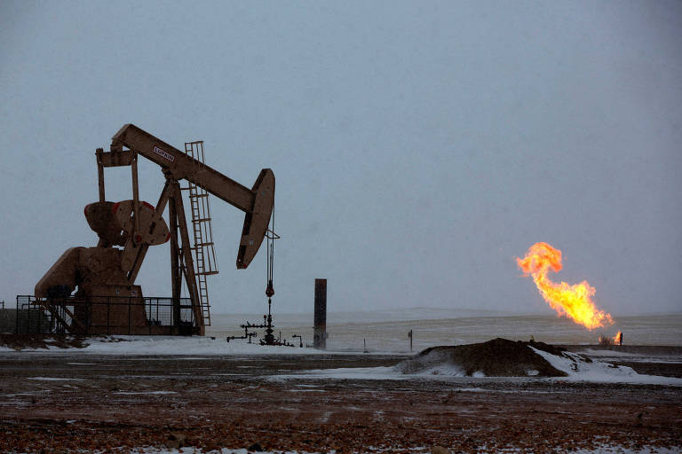 A imagem mostra uma bomba de extração de petróleo em operação em um campo coberto de neve. À direita, há uma chama de gás queimando, um flare de gás natural. O céu está nublado.