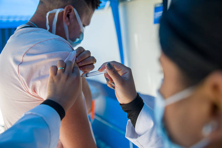 Profissional de saúde aplica vacina no braço de homem, que está de lado para ela, segurança a manga da camiseta 