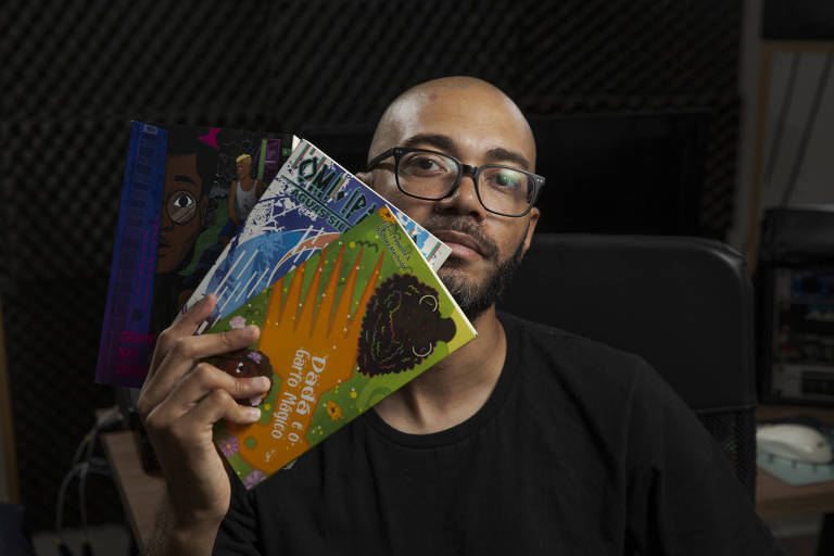 Retrato de Israel Neto, um homem negro, careca, com barba preta e curta; ele usa óculos de grau com armação preta e camiseta preta; com uma das mãos, ele segura três livros de sua editora, a Kitembo: 'Dadá e o Garfo Mágico', 'Omí Ìpalolo: Águas Silenciosas' e 'Crianças nas Sombras', todos com capas coloridas e ilustradas com personagens negros