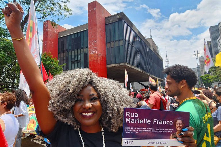 Mulher negra com cabelo grisalho e de punho erguido segura placa com os dizeres "rua Marielle Franco", com o Masp ao fundo
