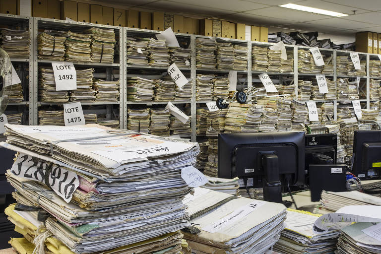 Pilhas de processos de papel sobre as mesas, monitores ao fundo e, atrás deles, uma ampla estante totalmente preenchida com mais pilhas de processos. Acima, várias caixas de arquivo de papelão