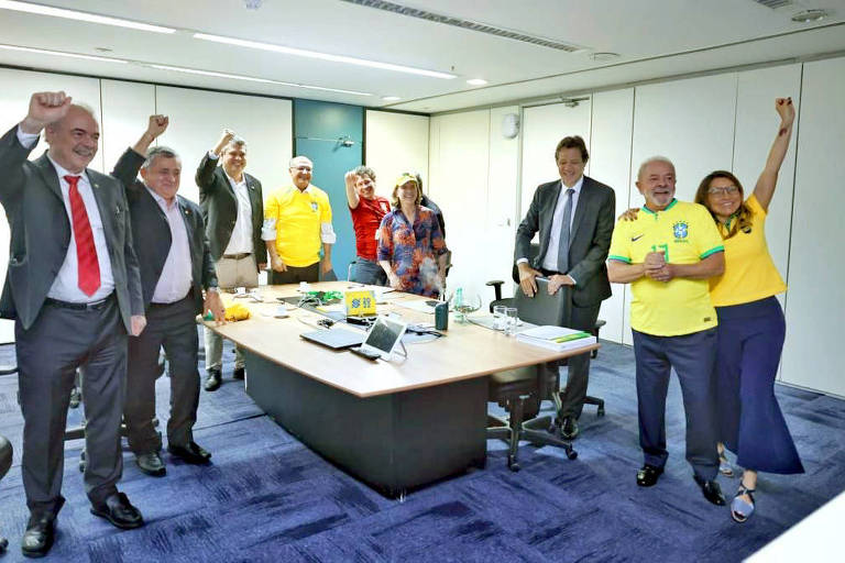 As nove pessoas aparecem em pé, vibrando, em uma sala com carpete azul e uma mesa ao centro